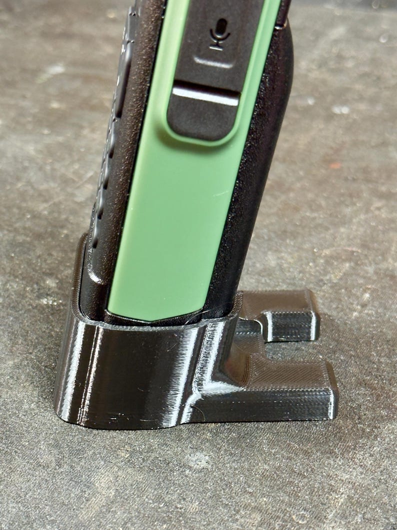 May include: A black 3D-printed cradle supporting a black and green handheld device. The cradle features a rectangular base with two supporting arms. The handheld device has a green side panel and a microphone icon.