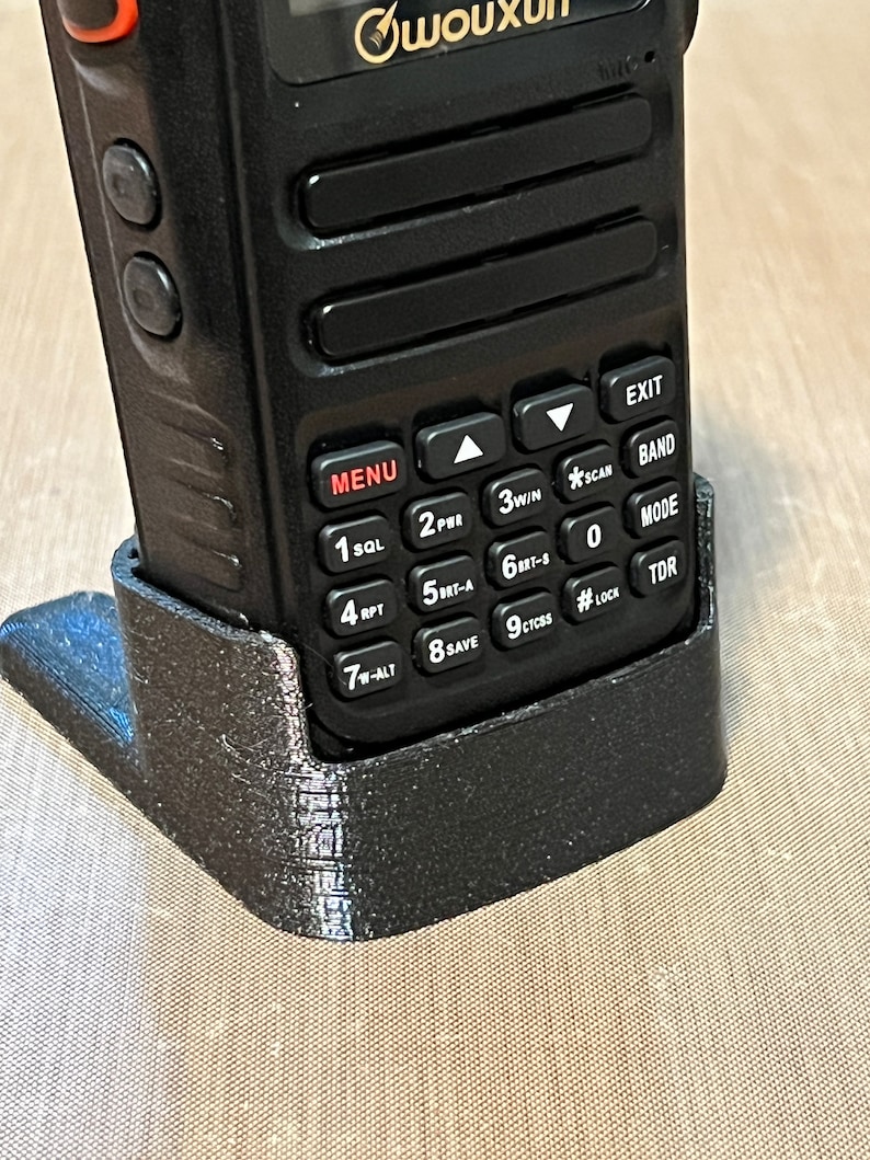 Pode incluir: Um r&aacute;dio bidirecional preto em um suporte preto. O r&aacute;dio possui um teclado com v&aacute;rios bot&otilde;es e uma tela. O suporte foi projetado para manter o r&aacute;dio em posi&ccedil;&atilde;o vertical. O r&aacute;dio tem a marca "Wouxun" impressa.