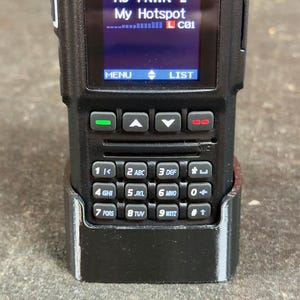 May include: A black handheld radio with a screen displaying the date and time, channel information, and the text "HS PNWR 2 My Hotspot L C01". The radio has a keypad with numbers and letters, and is in a black holster.