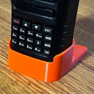 Pode incluir: Um r&aacute;dio port&aacute;til preto &eacute; mantido em um suporte laranja impresso em 3D. O r&aacute;dio possui um teclado com v&aacute;rios bot&otilde;es e uma tela. O suporte foi projetado para manter o r&aacute;dio em posi&ccedil;&atilde;o vertical. Os bot&otilde;es do r&aacute;dio incluem "MENU", "EXIT", "SCAN" e "MODE".