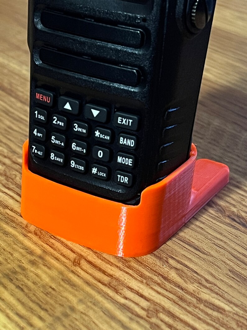 Pode incluir: Um r&aacute;dio port&aacute;til preto &eacute; mantido em um suporte laranja impresso em 3D. O r&aacute;dio possui um teclado com v&aacute;rios bot&otilde;es e uma tela. O suporte foi projetado para manter o r&aacute;dio em posi&ccedil;&atilde;o vertical. Os bot&otilde;es do r&aacute;dio incluem "MENU", "EXIT", "SCAN" e "MODE".