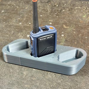 May include: A blue and black Rocky Talkie radio sits in a gray 3D-printed cradle. The radio has an orange button and antenna. The cradle has two additional compartments. The radio is designed for outdoor communication.