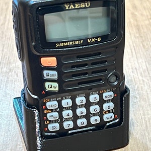 May include: A black Yaesu VX-6 submersible two-way radio with a keypad and a small LCD screen. The radio is in a black plastic holster.