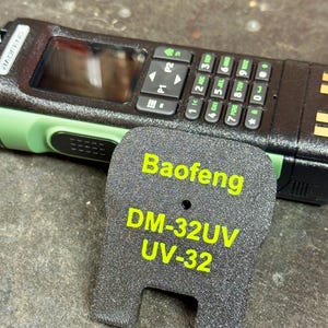 May include: A black and green Baofeng DM-32UV UV-32 radio with a keypad and screen. The radio is next to a black accessory with the Baofeng model name and number in yellow text. The radio has a green accent.