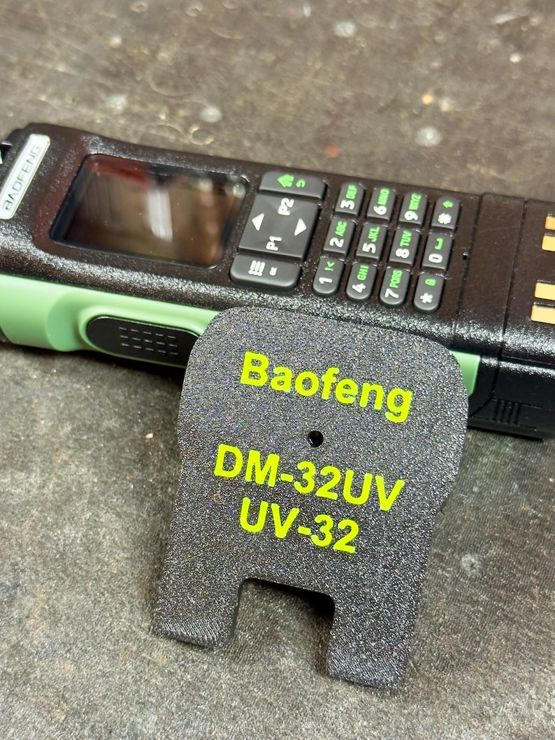 May include: A black and green Baofeng DM-32UV UV-32 radio with a keypad and screen. The radio is next to a black accessory with the Baofeng model name and number in yellow text. The radio has a green accent.