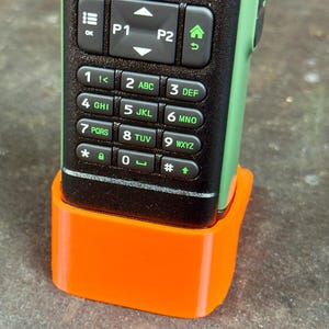 May include: A black and green handheld radio with an orange base. The radio has a keypad with numbers and letters, and several function buttons. The base is a bright orange colour and appears to be 3D printed.