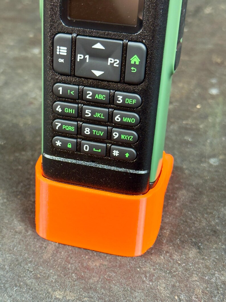 May include: A black and green handheld radio with an orange base. The radio has a keypad with numbers and letters, and several function buttons. The base is a bright orange colour and appears to be 3D printed.