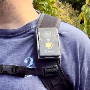 May include: A black and gray Sensecap device attached to a black backpack strap. The device has a clear top with internal components visible, a yellow button, and the Sensecap logo. The strap is secured with a black plastic buckle. The person is wearing a blue shirt.
