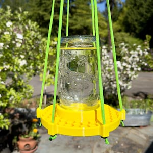 May include: A clear glass jar with embossed fruit designs sits on a bright yellow circular plastic base with a textured surface. The assembly is suspended by four neon green nylon cords attached to the base, creating a hanging feeder structure.