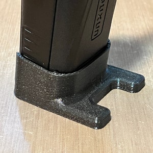 Pode incluir: Um objeto preto retangular com a palavra "UNXUN" impressa na lateral, repousando em um suporte preto texturizado feito sob medida. O suporte tem uma base em forma de T para estabilidade.