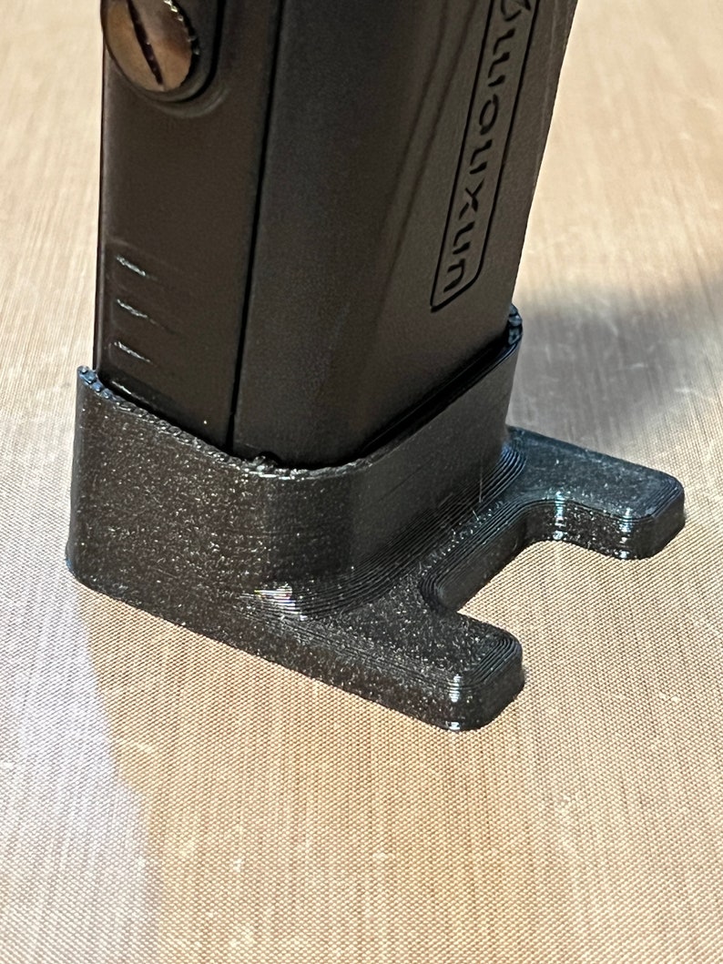 Pode incluir: Um objeto preto retangular com a palavra "UNXUN" impressa na lateral, repousando em um suporte preto texturizado feito sob medida. O suporte tem uma base em forma de T para estabilidade.