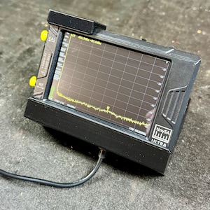 May include: A black electronic device with a screen displaying a yellow line graph. The device has yellow knobs and the text "tinySA ULTRA". A black cable is connected to the device.