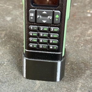 May include: Close-up of a two-way radio with a black keypad and green accents. The keypad has numbers, letters, and function buttons. The radio is in a black base.
