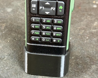May include: Close-up of a two-way radio with a black keypad and green accents. The keypad has numbers, letters, and function buttons. The radio is in a black base.