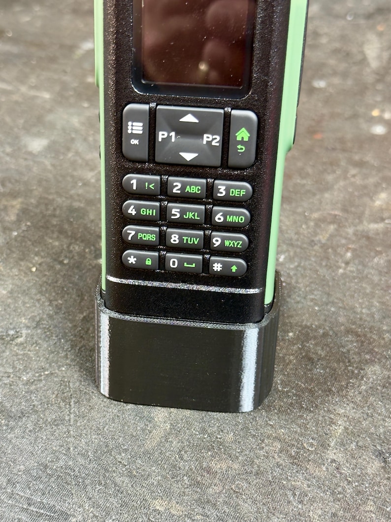 May include: Close-up of a two-way radio with a black keypad and green accents. The keypad has numbers, letters, and function buttons. The radio is in a black base.