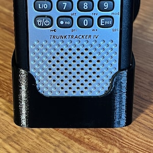 Puede incluir: Un escáner de mano plateado y negro con una funda negra. El escáner tiene un teclado con botones etiquetados 'Scan', 'srch', 'L/0', 'att', 'no', 'pri', 'wx', 'gps', 'Eyes', 'mod', 'disp', 'if', 'lvl', 'sr1', 'sr2', 'sr3', '4', '5', '6', '7', '8', '9', '0', '<', '>'. El escáner también tiene una rejilla de altavoz y el texto 'TRUNKTRACKER IV'.