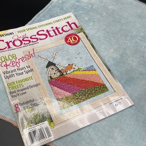 May include: A magazine cover for Just Cross Stitch, a cross-stitch magazine. The cover features a colorful cross-stitch design of a windmill and flowers. The magazine is celebrating its 40th anniversary.