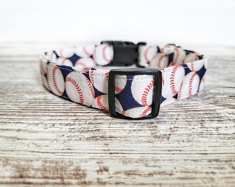 Puede incluir: Un collar de perro blanco y azul marino con un patrón de béisbol. El collar tiene una hebilla de plástico negro y un anillo en D de metal negro.