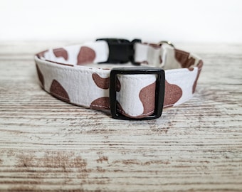 May include: A white and brown dog collar with a black buckle. The collar has a brown cow print pattern.