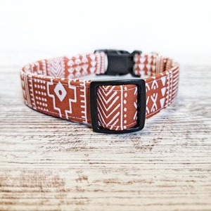May include: A dog collar with a geometric pattern in shades of brown and white. The collar has a black plastic buckle and a matching black square loop. The fabric features repeating patterns of shapes and lines.