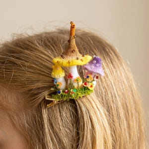 May include: A whimsical hair clip featuring a cluster of colorful mushroom figures. The clip showcases a large, tan and yellow mushroom with a beaded stem, and smaller yellow and purple mushrooms. The mushrooms are set on a bed of green moss with tiny flowers.