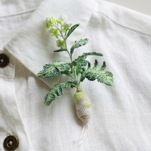 Puede incluir: Broche de rábano de ganchillo hecho a mano con hojas verdes y raíz blanca y amarilla, prendido a una camisa de lino blanca. El broche tiene una apariencia detallada y texturizada, mostrando el arte de la artesanía. El rábano es un accesorio único.