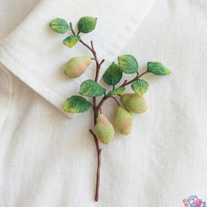 May include: A handmade brooch depicting a branch with green leaves and small, pear-shaped fruits. The fruits display a gradient of green and pink tones. The brooch is set against a white fabric background.