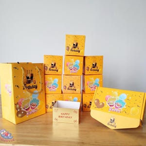 May include: A collection of yellow birthday gift boxes and a gift bag. The boxes and bag feature the words "Happy Birthday" and cartoon characters. The boxes are stacked and arranged on a wooden surface.