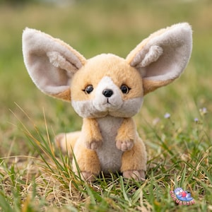 May include: A plush Fennec fox toy with large, beige-lined ears, a tan body, and a white face. The stuffed animal is sitting in green grass, with a soft, cuddly appearance. The toy has dark eyes and a small tail.