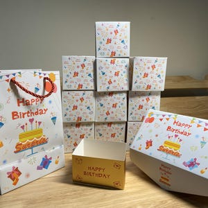 May include: A selection of birthday gift packaging, including a white gift bag with red handles and various boxes. The bag and boxes are decorated with colourful birthday graphics, such as a cake, balloons, and the words "Happy Birthday".