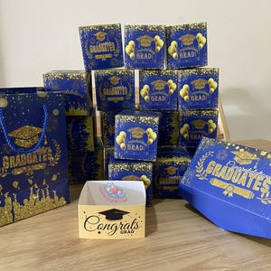 May include: A collection of graduation-themed gift boxes and a gift bag. The boxes are navy blue with gold detailing, featuring graduation caps and the words "GRAD" and "GRADUATES". The gift bag is navy blue with gold glitter and the word "GRADUATES".