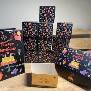 Peut inclure: Ensemble de boîtes et sacs cadeaux sur le thème de l'anniversaire. Les boîtes et sacs noirs présentent des illustrations colorées de feux d'artifice, de gâteaux et de cadeaux. Le texte "Happy Birthday" et "Surprise" est visible.