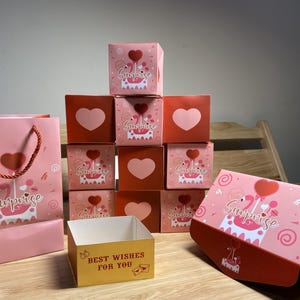 Può includere: Una collezione di scatole regalo rosa e rosse e una borsa regalo. Le scatole presentano disegni a cuore e torta, con la parola "Surprise" su alcune. È presente anche una scatola gialla con la scritta "Best Wishes For You".