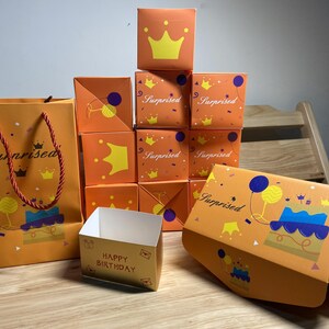 Puede incluir: Cajas de regalo y una bolsa de regalo de color naranja con diseños de cumpleaños. Las cajas presentan coronas y la palabra "Surprised". La bolsa de regalo y una caja muestran un pastel y globos. Las palabras "Happy Birthday" están en una caja pequeña.