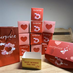 Op de afbeelding: Een verzameling rode en roze geschenkdozen en een cadeauzak. De dozen hebben hartontwerpen en het woord "verrassing". De cadeauzak heeft een hart- en ballonnenontwerp en het woord "verrassing". Een kleine gouden doos zegt "Beste wensen voor jou".