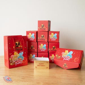 May include: A selection of red gift boxes and a gift bag, all decorated with a birthday theme. The boxes and bag display the words "Happy Birthday" and cartoon characters with balloons. A small gold box with "Happy Birthday" is also visible.