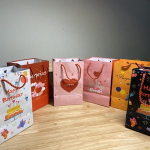 Può includere: Una collezione di sacchetti regalo colorati con manici in corda ritorta. Le borse presentano temi di compleanno e sorpresa, con disegni di torte e cuori. I colori includono bianco, arancione, rosa, giallo e nero.