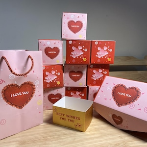 May include: A selection of pink and red gift boxes and a gift bag. The boxes display heart designs with the text "I LOVE YOU" and "BEST WISHES FOR YOU". The bag has a heart design and a twisted rope handle.
