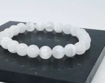 Può includere: Un braccialetto di perline bianche. Il braccialetto è composto da perline bianche rotonde e opache. Le perline sono infilate insieme, con una superficie liscia e dimensioni uniformi. Il braccialetto è esposto su una superficie scura.