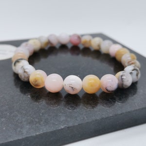 Könnte beinhalten: Ein Armband aus kugelförmigen Perlen in Rosa-, Weiß- und Gelbtönen, wobei einige Perlen graue und schwarze Markierungen aufweisen. Das Armband wird auf einer dunkelgrauen Oberfläche präsentiert.