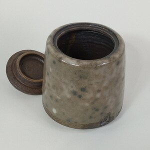 Op de afbeelding: Een kleine, gevlekte keramische pot met een deksel. De pot is lichtbruin van kleur met donkerbruine vlekken. Het deksel is donkerbruin.