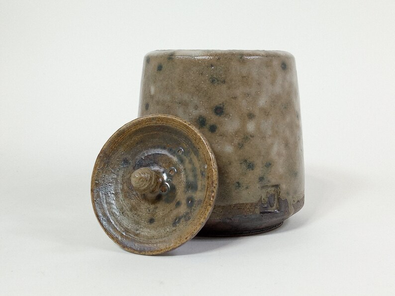 Op de afbeelding: Een keramische pot met een deksel, beide met een gespikkeld bruin en wit glazuur. Het deksel heeft een kleine knop bovenop.