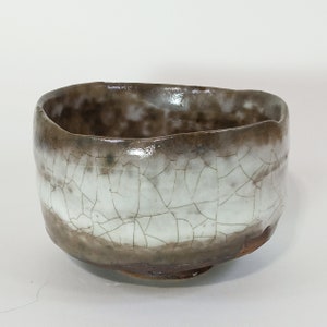 May include: A white ceramic tea bowl with a brown glaze and a crackled finish. The bowl has a rounded shape and a slightly flared rim.