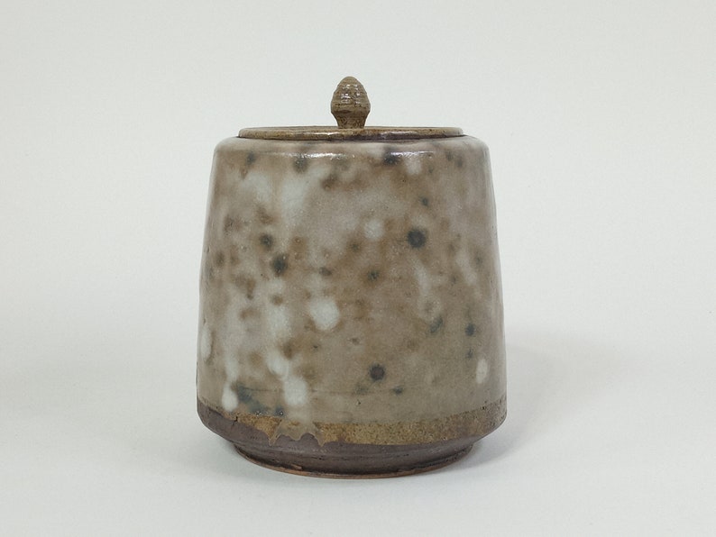 Op de afbeelding: Een keramische pot met een deksel, met een gespikkeld bruin en wit glazuur. De pot heeft een ronde basis en een licht uitlopende opening.