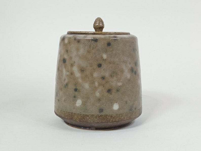 Op de afbeelding: Een keramische pot met een gespikkelde bruine en witte glazuur en een houten deksel. De pot heeft een ronde basis en een licht uitlopende bovenkant.