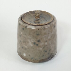 Op de afbeelding: Een kleine, gevlekte keramische pot met een deksel. De pot is lichtbruin van kleur met donkerbruine vlekken. Het deksel heeft een klein knopje bovenop.