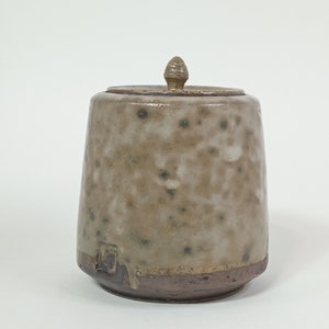 Op de afbeelding: Een keramische pot met een deksel en een gespikkelde glazuur. De pot is lichtbruin van kleur met donkerbruine spikkels. Het deksel heeft een klein knopje bovenop.