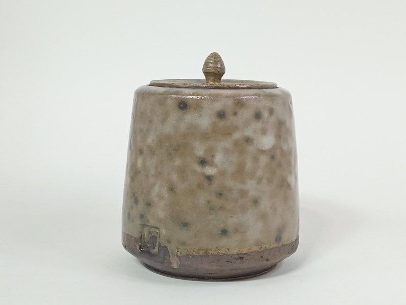 Op de afbeelding: Een keramische pot met een deksel en een gespikkelde glazuur. De pot is lichtbruin van kleur met donkerbruine spikkels. Het deksel heeft een klein knopje bovenop.