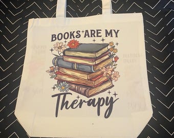 May include: Off-white canvas tote bag with white handles. The bag features a graphic of a stack of books with floral accents and the text "BOOKS ARE MY Therapy" in black script. The background is a black and white chevron pattern.