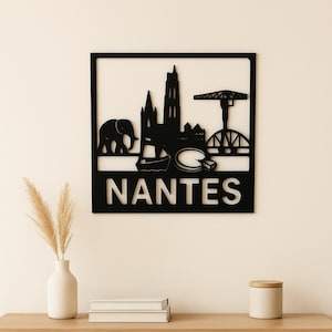 Peut inclure: Décoration murale en métal noir représentant la silhouette de Nantes, avec un éléphant, une cathédrale et le nom de la ville. L'œuvre est exposée au-dessus d'un meuble en bois avec un vase, des livres et un coussin décoratif.